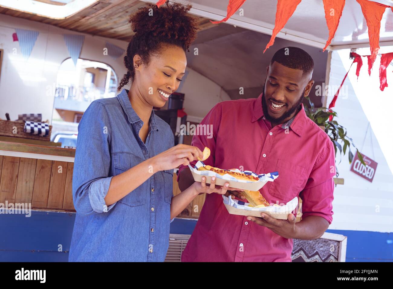 Lächelndes, vielfältiges Paar, das Hot Dogs und Kartoffelkeile isst Foodtruck an sonnigen Tagen Stockfoto