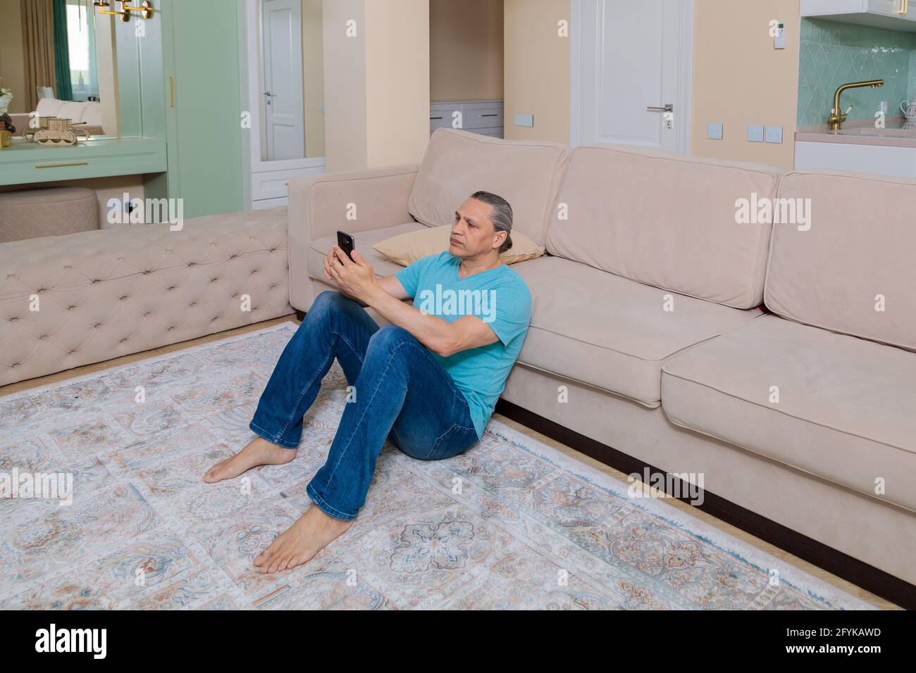 Ein erwachsener Mann durchsucht soziale Netzwerke auf einem Mobiltelefon, während er in einer Wohnung auf dem Boden sitzt. Stockfoto