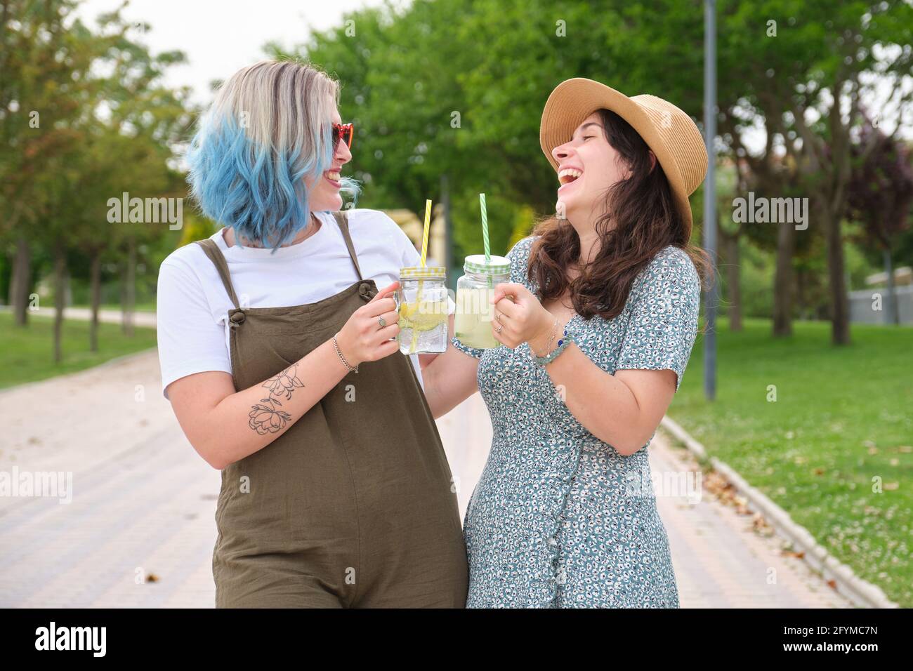 Zwei glückliche Freundinnen trinken erfrischende Getränke, lachen und schauen sich an einem sonnigen Sommertag in einem Park an. Stockfoto