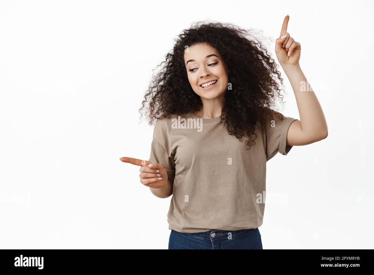 Schöne lockige Mädchen zeigt Finger seitwärts, zwei Möglichkeiten, entscheiden, links Produkt zu wählen, stehend in T-Shirt vor weißem Hintergrund, Einkaufen in Stockfoto
