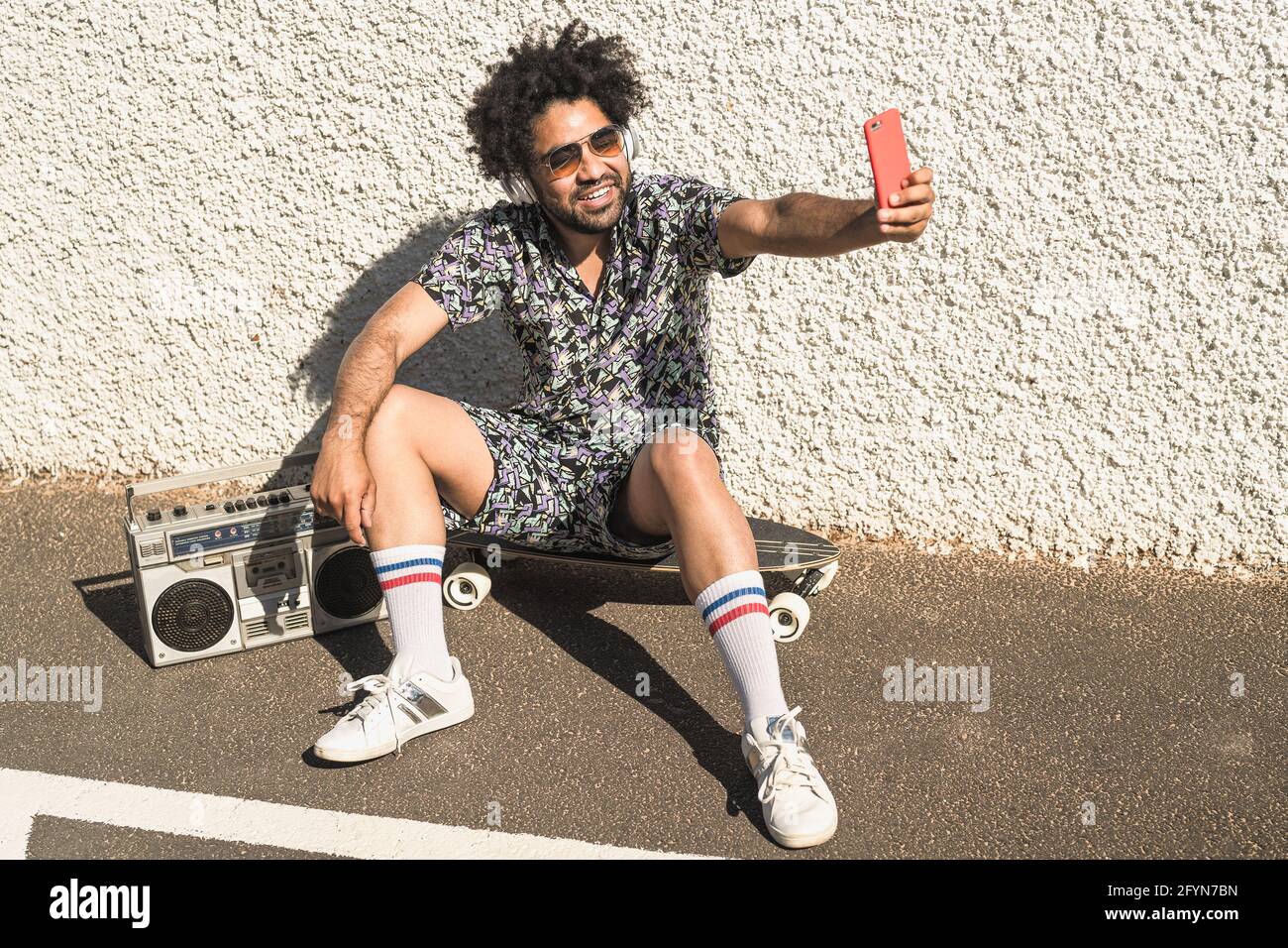Junger afro-lateinischer Mann, der Spaß beim Selfie mit dem Handy hat Smartphone, während Sie Musik mit Kopfhörern und Vintage-Boombox hören Sommerferien Stockfoto