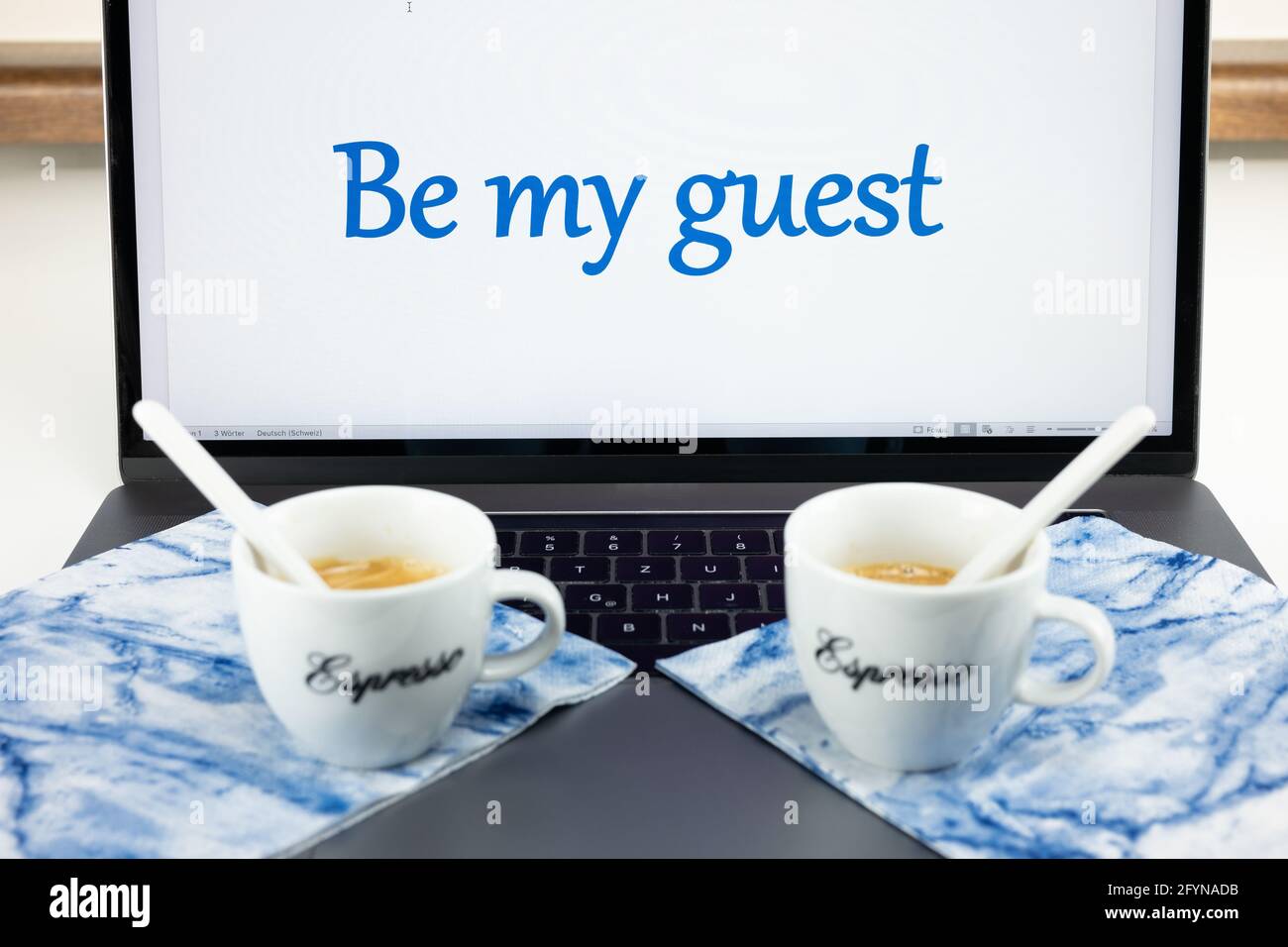 Zwei Kaffeetassen liegen auf einem Desktop-Computer, und auf dem Bildschirm steht, dass ich mein Gast sein soll. Stockfoto