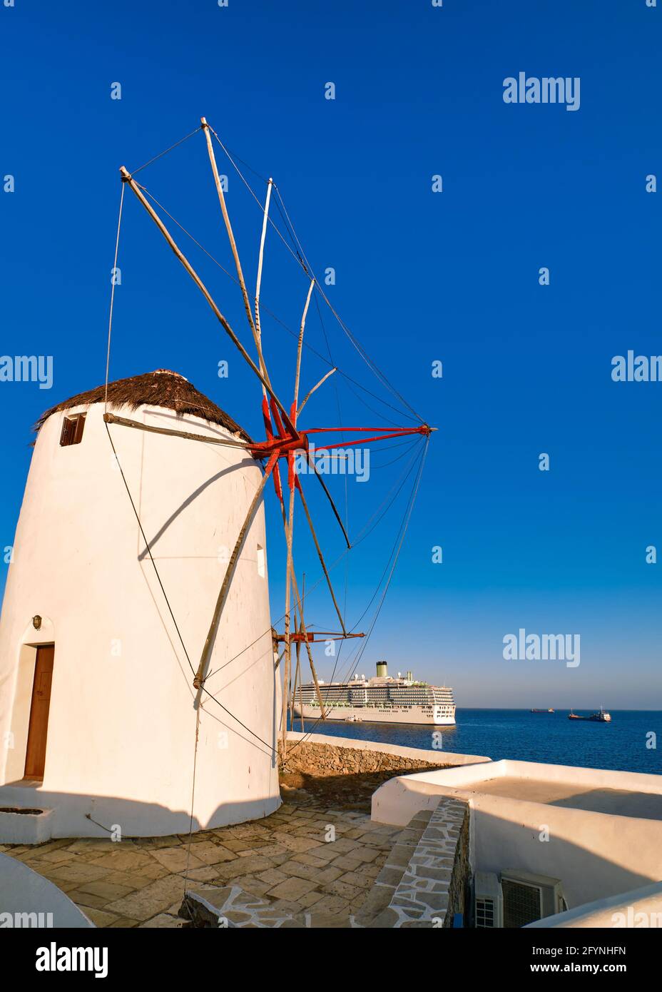 Berühmte Touristenattraktion von Mykonos, Griechenland. Traditionelle, weiß getünchte Windmühle auf dem Seeweg und Kreuzfahrtdampfer, der den Hafen verlässt. Morgen, Sommer, klarer blauer Himmel Stockfoto