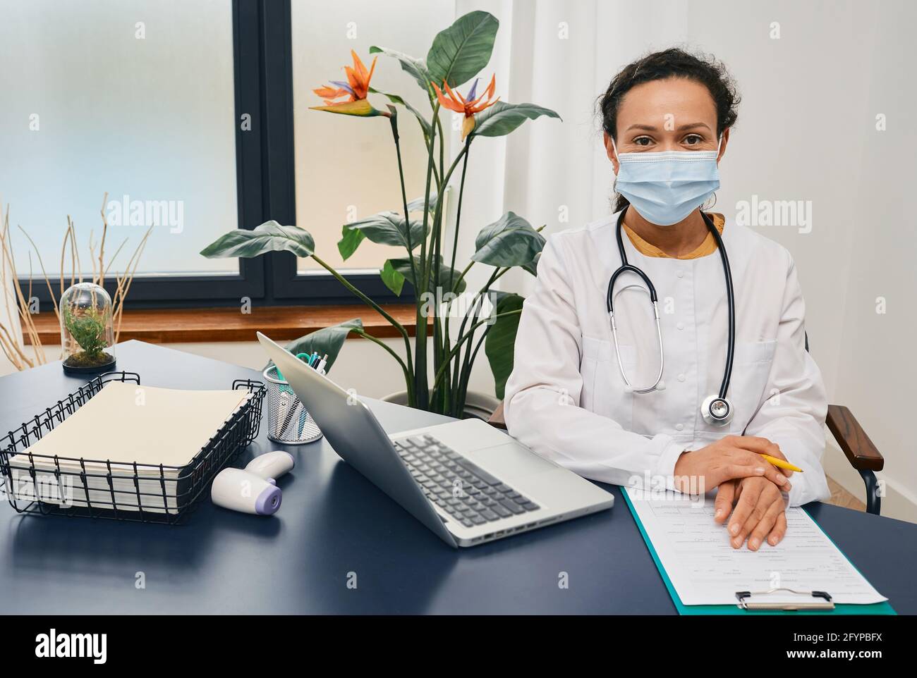 Porträt einer Hausarztfrau mit Schutzmaske und Arztkittel, der in ihrem Arbeitsbüro bei einem Arzt sitzt Klinik Stockfoto