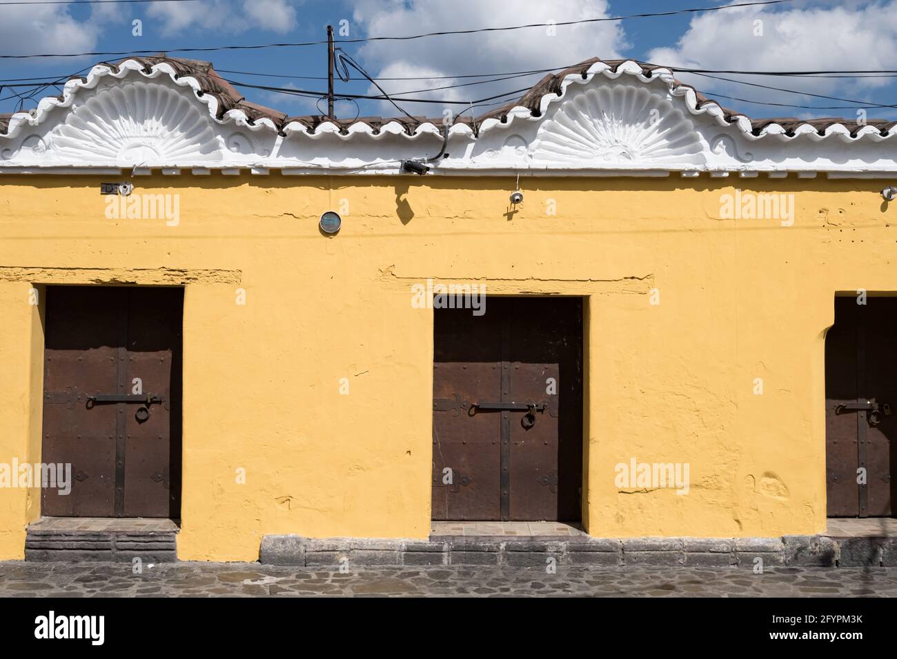 Detail der Fassade eines Gebäudes in Antigua Guatemala mit verzierten Dachstilen, lackierten gelben Wänden und eisernen Türen. Stockfoto
