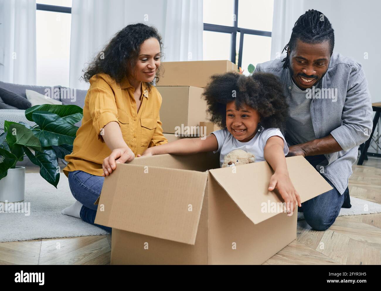Nettes afroamerikanisches kleines Mädchen, das in einer Kartonbox sitzt und mit ihren Eltern spielt, während es in ein neues Zuhause zieht Stockfoto
