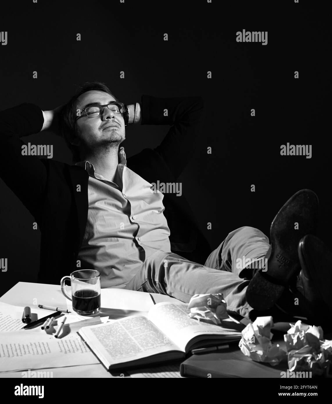 Der Erwachsene Mann in legerer Kleidung und Brille sitzt in verträumter Pose mit den Händen hinter dem Kopf, den Füßen auf dem Tisch mit Buch und Papieren Stockfoto