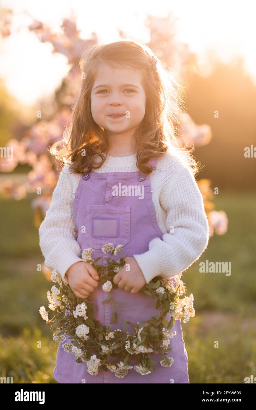 Nettes Kind Mädchen 2-3 Jahre alt halten Blumenkranz über blühenden Baum und Sonnenuntergang im Park im Freien. Tragen Sie legere Kleidung über dem Hintergrund der Natur. Kindheit Stockfoto