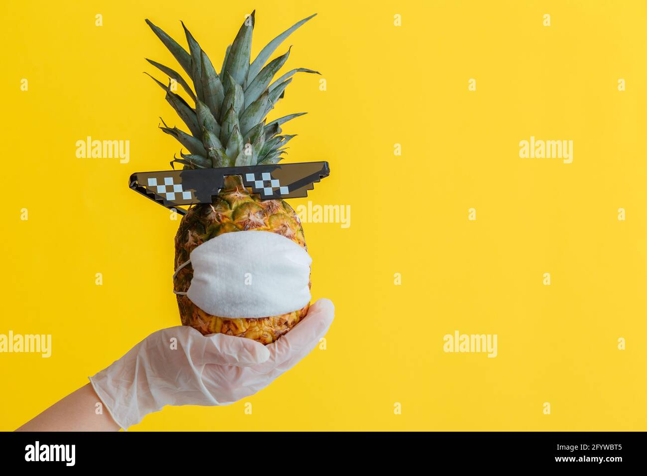 Hand in Hand halten Ananas in medizinischer Gesichtsmaske und Sonnenbrille auf gelbem Hintergrund. Ananasfrucht ist im Covid Coronavirus-Lockdown zu sehen Stockfoto