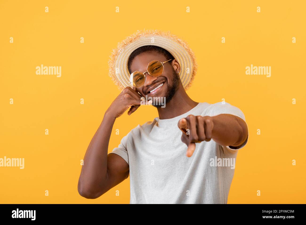 Fröhlicher schwarzer Kerl in trendigem Sommeroutfit, der Call Me-Geste zeigt und auf gelbem Studiohintergrund die Kamera anlächelt Stockfoto