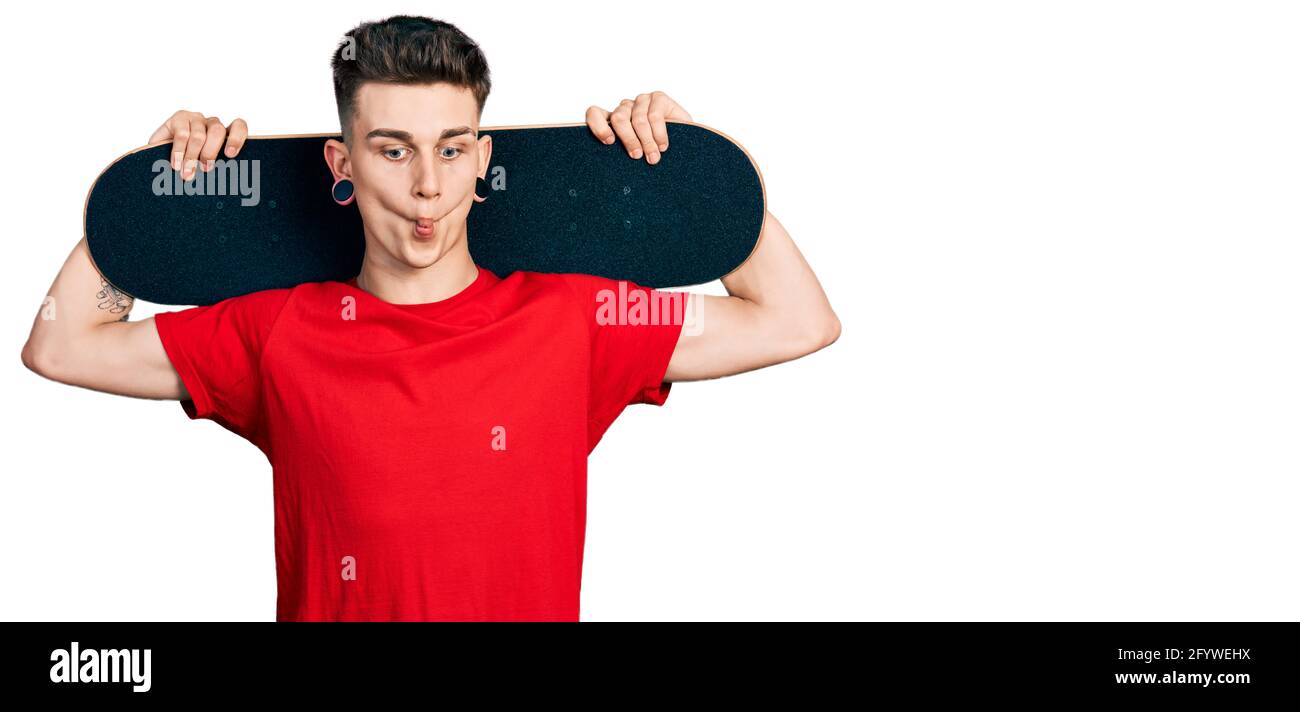 Junge kaukasische Junge mit Ohren Erweiterung halten Skate über Schultern machen Fische Gesicht mit Mund und schielende Augen, verrückt und komisch. Stockfoto