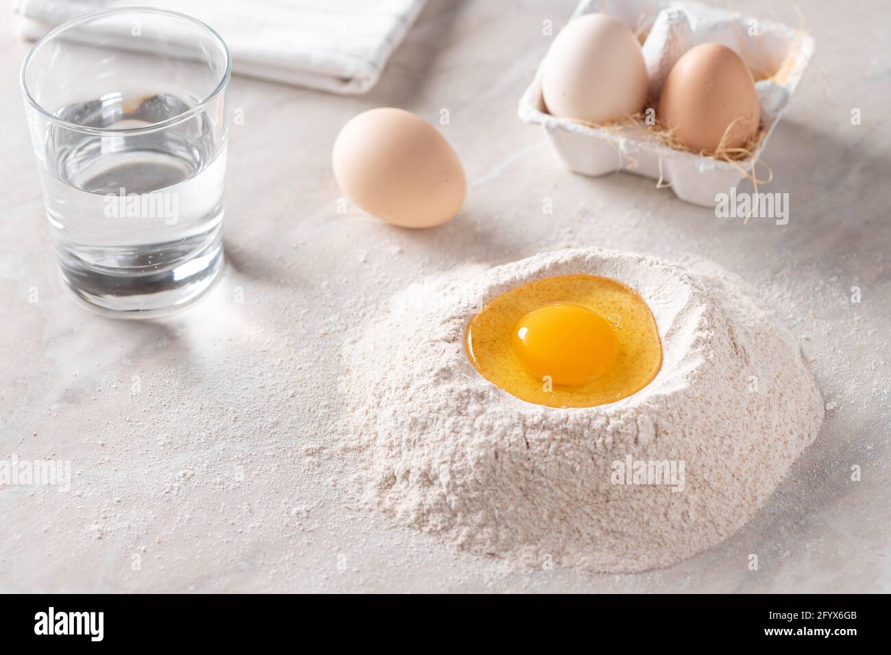 Haufen Vollkornmehl mit rohem Ei in der Mitte, Glas Wasser und Eier auf dem Küchentisch Stockfoto