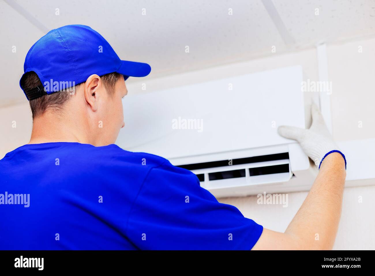 Ein netter Techniker mit blauer Baseballkappe und Overalls fixiert die neue Klimaanlage an der Wand im Zimmer. Porträt eines Arbeiters von hinten. Modell Stockfoto