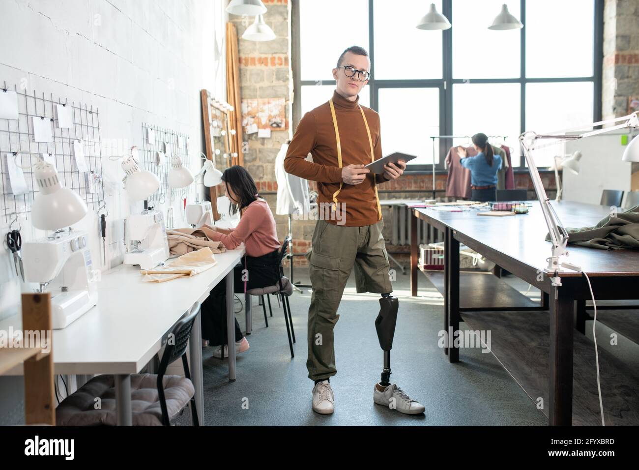 Junge Disable Schneider stehen zwischen Tisch und Nähmaschinen in Große Werkstatt Stockfoto