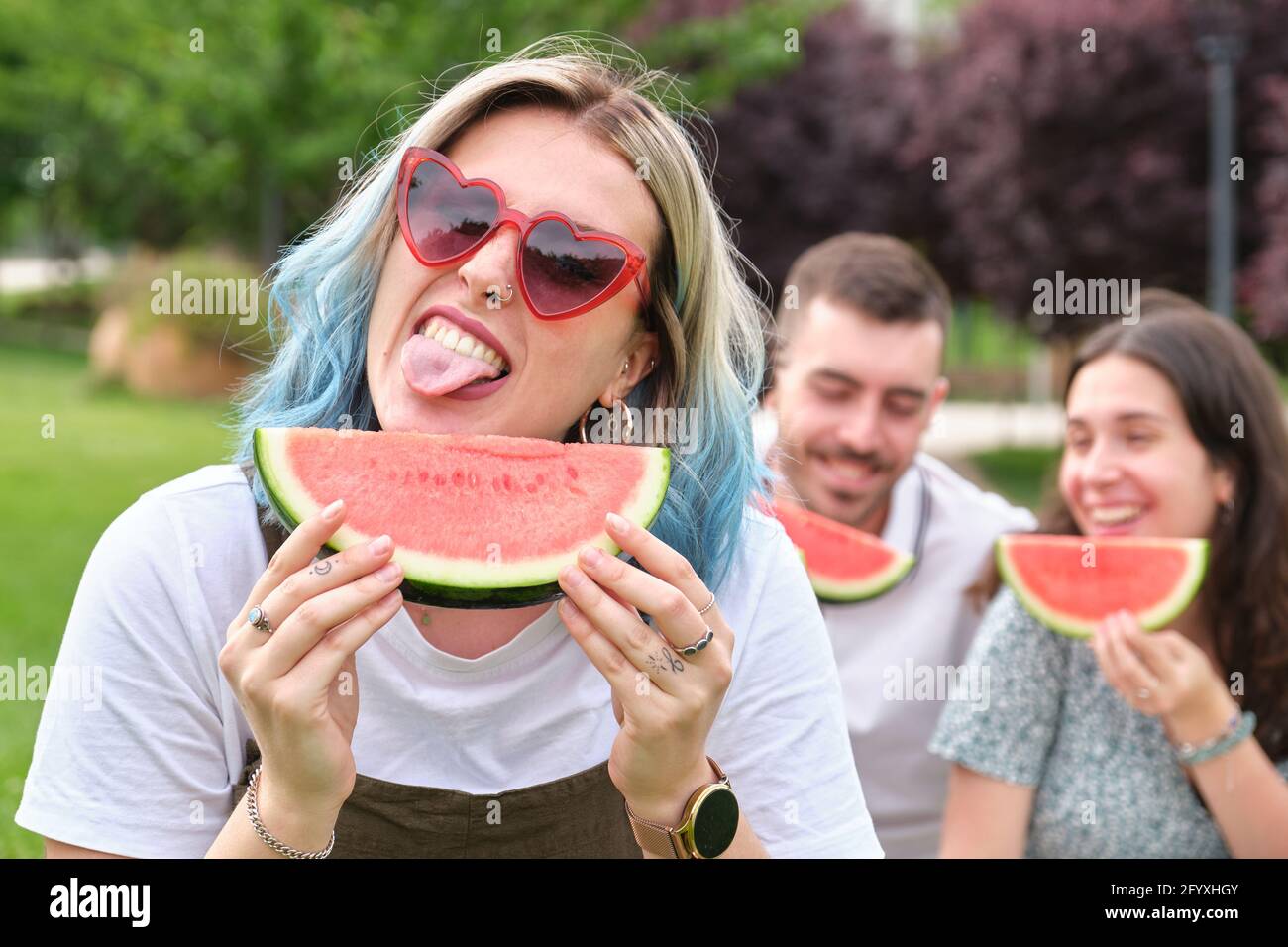 Glückliche junge Frau, die ihre Zunge herausstreckt und ein Stück  Wassermelone hält, und Freunde lächeln auf dem Hintergrund Stockfotografie  - Alamy