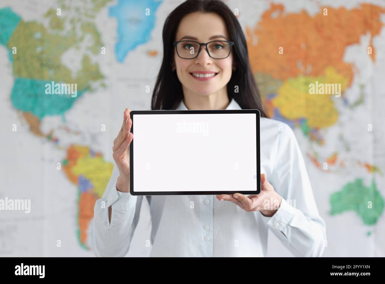 Lächelnde Frau in einer Brille hält ein Tablet mit weißem, leeren Bildschirm Auf dem Hintergrund der globalen Weltkarte Stockfoto