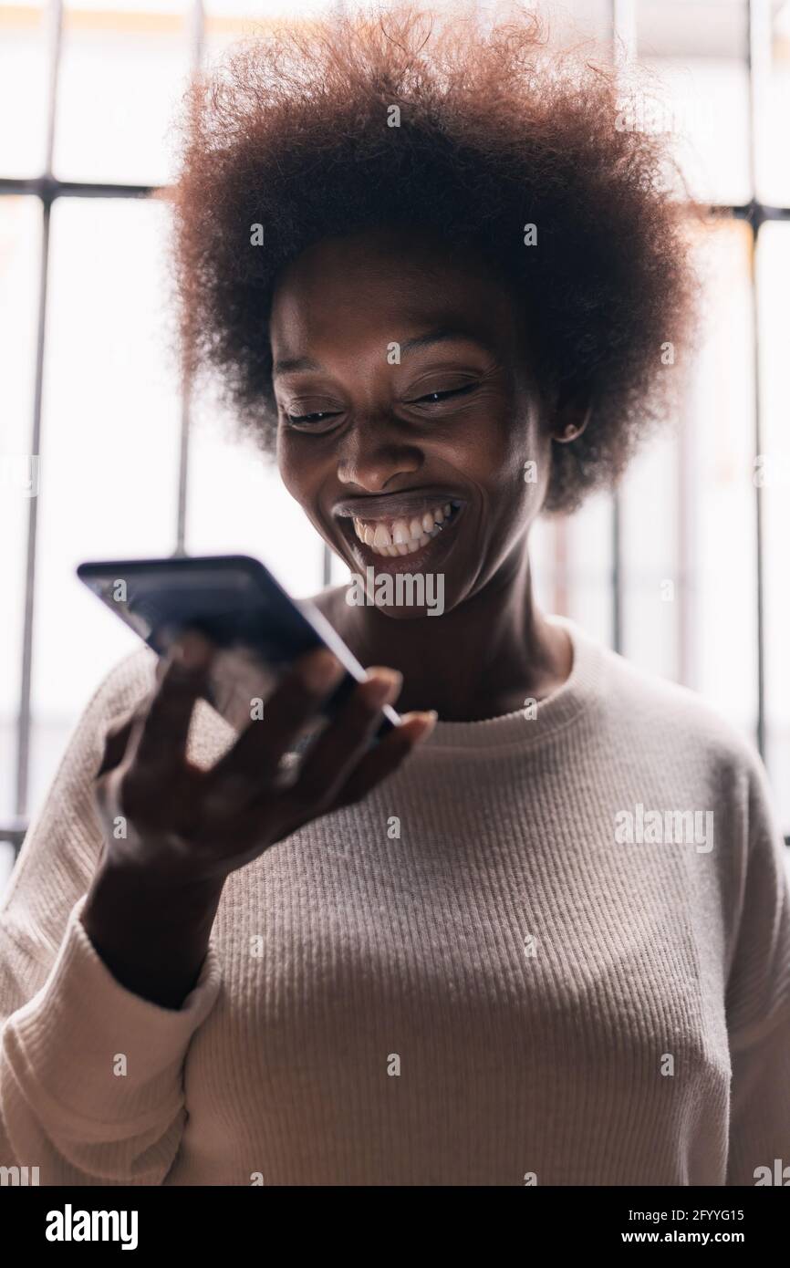Fröhliche junge afroamerikanische Frau mit lockigen dunklen Haaren lächelnd Und Sprachnachrichten zu Hause auf dem Mobiltelefon aufzeichnen Stockfoto
