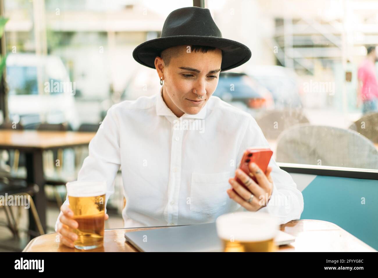 Konzentrierter Tomboy in Hut sitzend am Tisch mit Laptop und Ein Glas Bier beim Scrollen auf dem Mobiltelefon Stockfoto