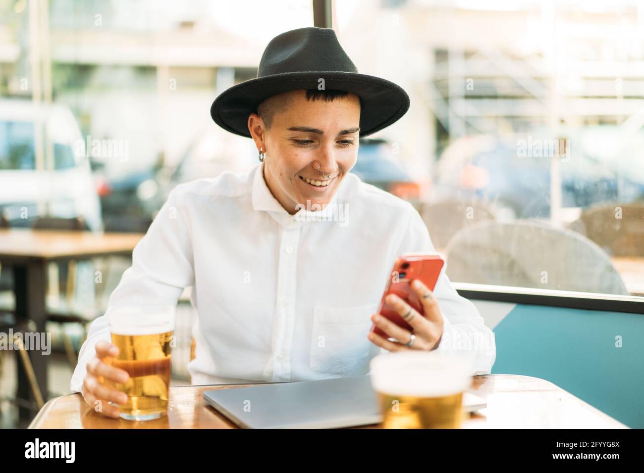 Konzentrierter Tomboy in Hut sitzend am Tisch mit Laptop und Ein Glas Bier beim Scrollen auf dem Mobiltelefon Stockfoto