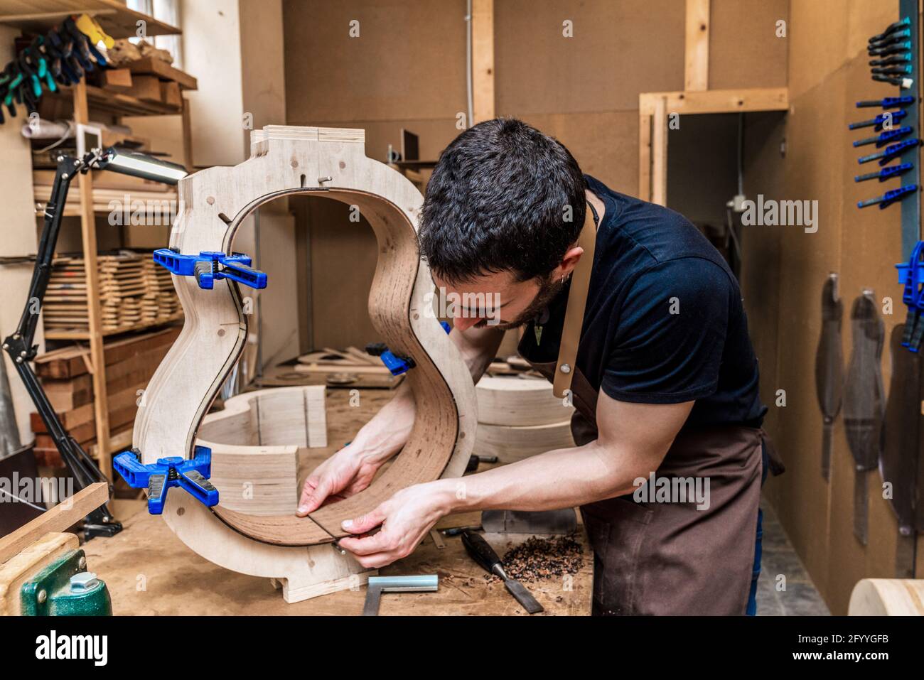 Männlicher Handwerker, der sich nach vorne lehnt, während er ein hölzernes Musikinstrument repariert Mit Klemmen im Arbeitsraum mit professionellen Werkzeugen Stockfoto