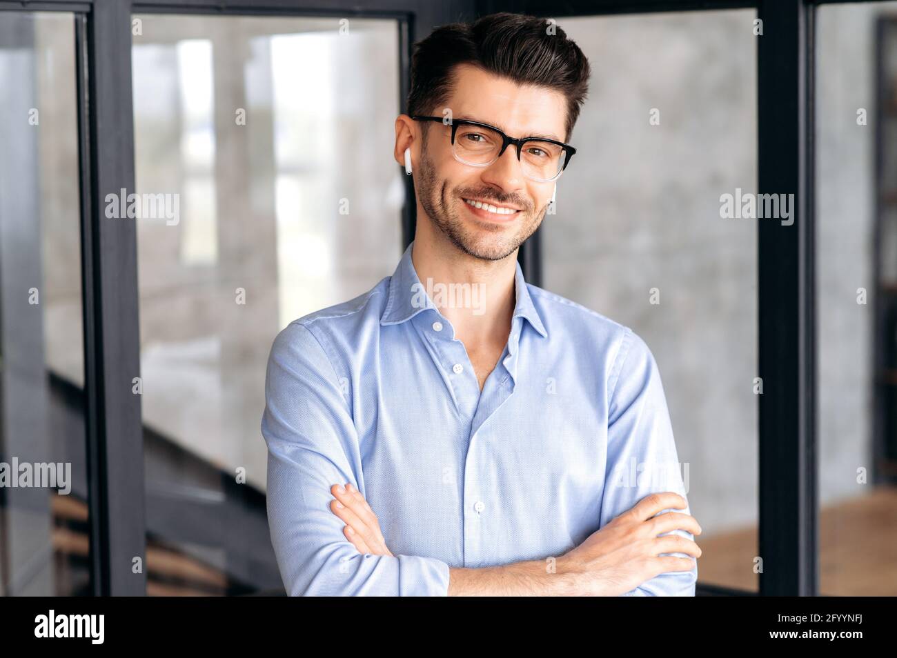 Porträt eines fröhlichen, attraktiven, selbstbewussten jungen Geschäftsmanns oder Geschäftsführers, der Brille und Kopfhörer trägt, ein formelles Hemd trägt, im Büro steht, die Kamera anschaut, die Arme gekreuzt, Freundlich lächelnd Stockfoto