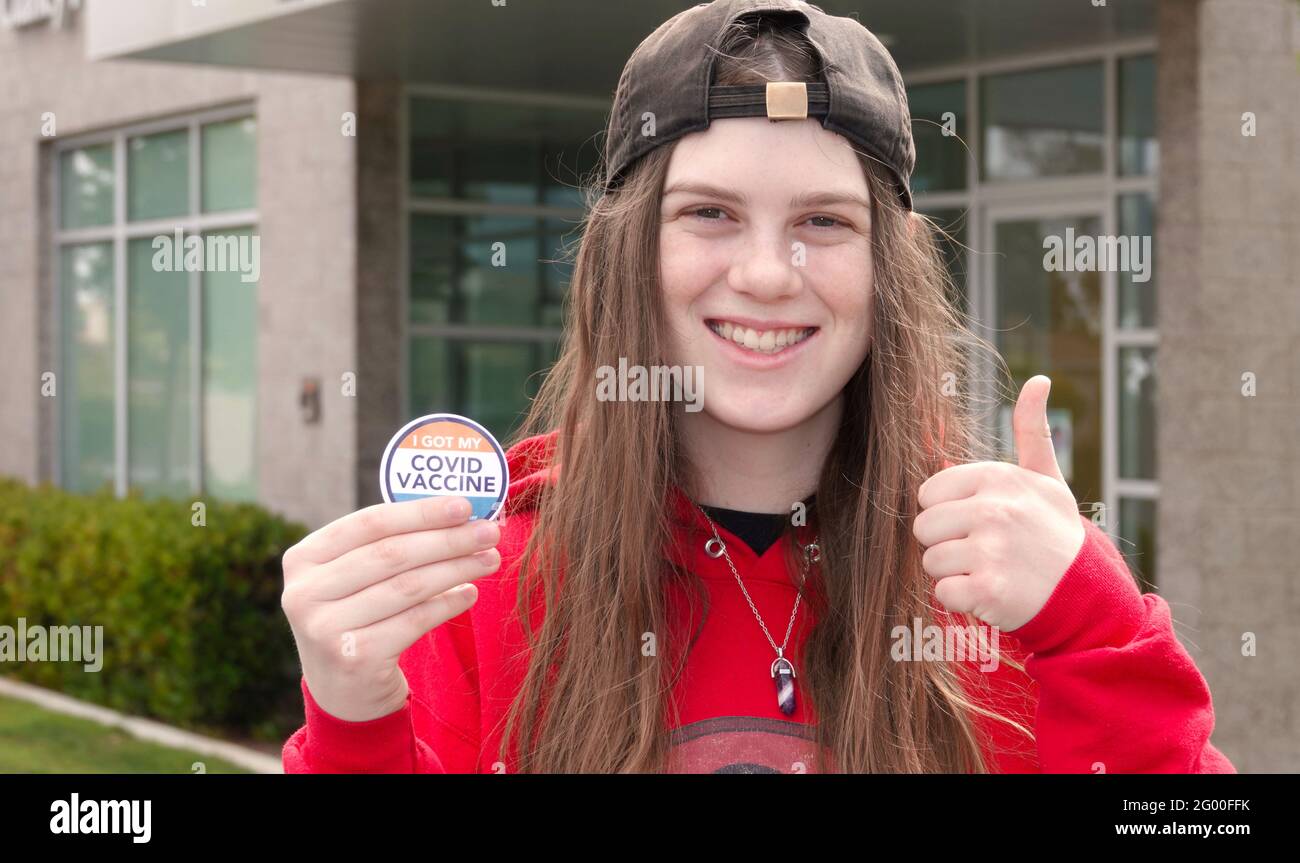 Ein glücklicher, geimpfter Teenager lächelt und gibt einen Daumen nach oben Während sie ihren Covid-19 Impfstoffaufkleber in der Hand hält Stockfoto