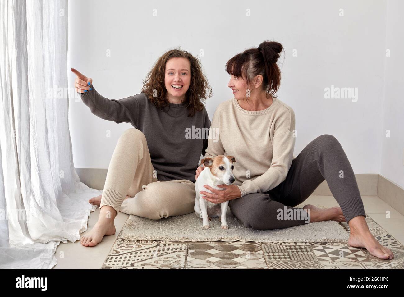 Fröhliche Erwachsene Tochter und Mutter mittleren Alters sitzen auf dem Boden Mit niedlichem Hund zu Hause Stockfoto