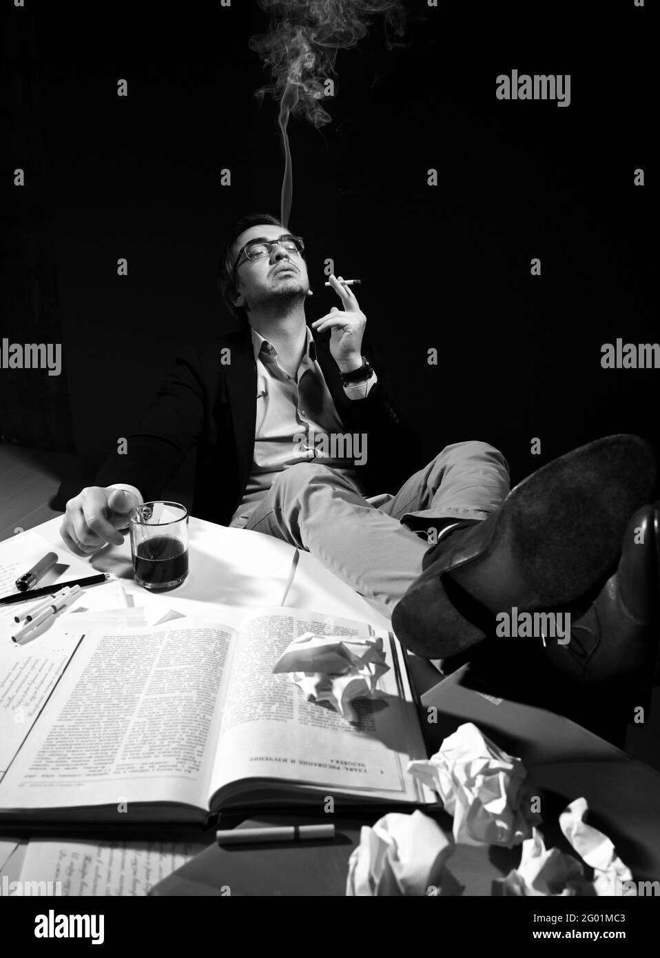 Der Erwachsene Mann in legerer Kleidung und Brille sitzt mit den Füßen auf dem Tisch mit Büchern und Papieren, trinkt Tee, Kaffee und raucht Stockfoto
