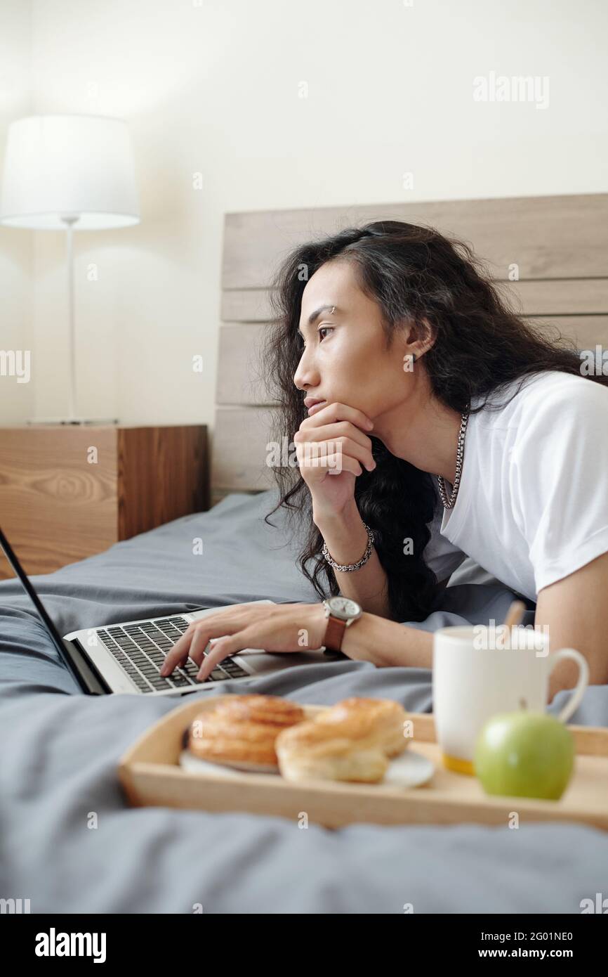 Nachdenklicher junger vietnamesischer Online-Manager mit langen Haaren Übernachtung mit Frühstück auf dem Tablett und mit einem Laptop beim Nachdenken Des Projekts Stockfoto