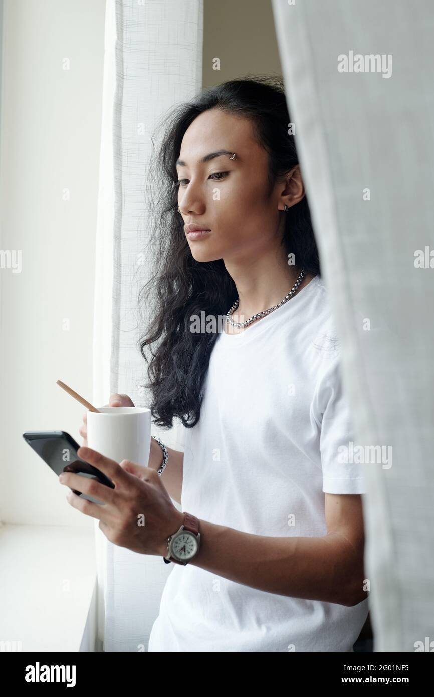 Seriöser junger Vietnamese mit welligen Haaren, die am Fenster stehen Und Kaffee trinken, während Sie durch die sozialen Medien blättern Stockfoto