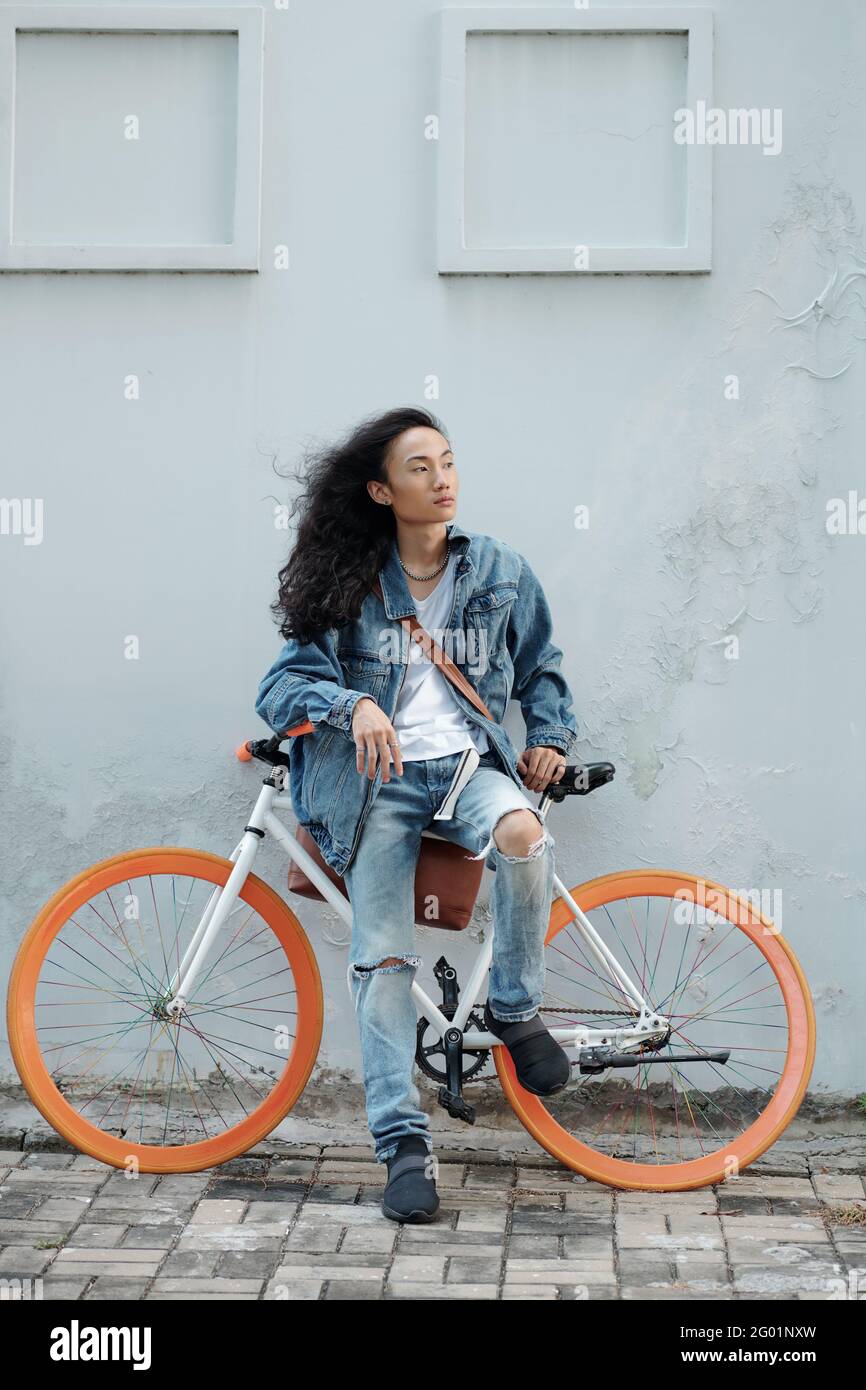 Serious hübsch junge vietnamesische Modell mit langen Haaren im Wind Sitzen auf dem Fahrrad mit orangefarbenen Rädern und Blick in die Ferne Stockfoto