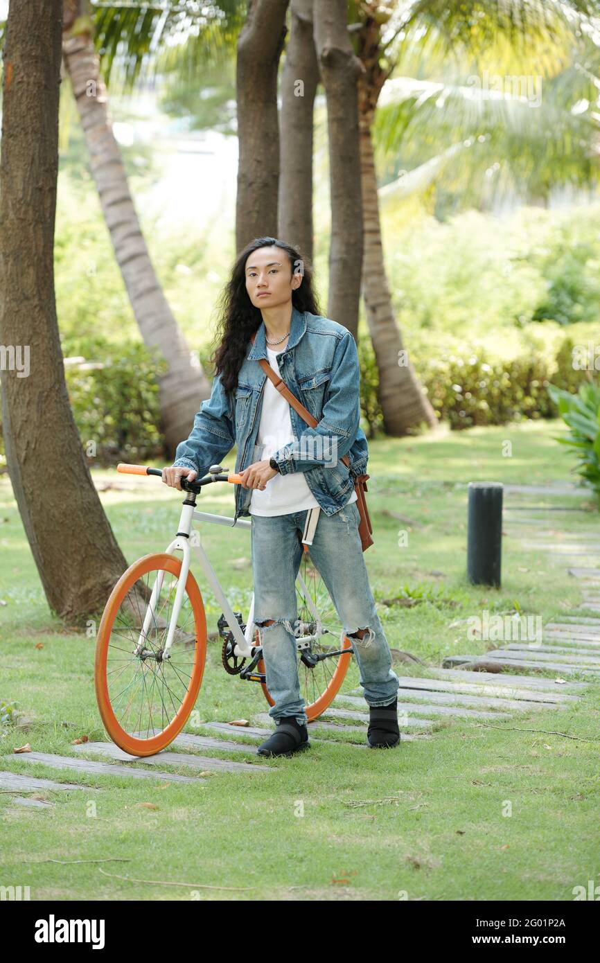 Serious junge Vietnamese Kerl in Denim tragen zu Fuß mit Fahrrad Auf dem Weg in einem tropischen Park mit Palmen Stockfoto