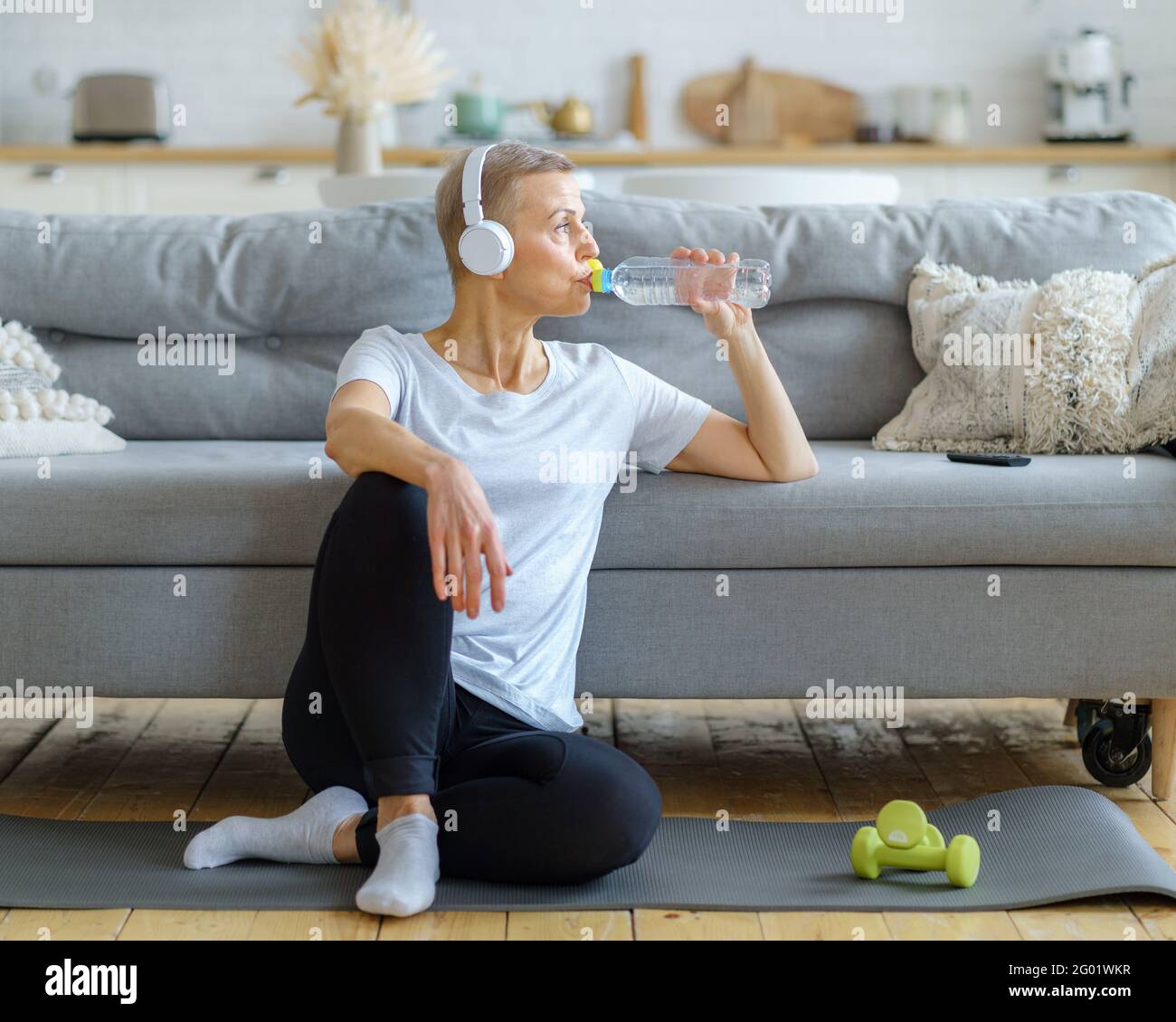 Glückliche ältere Frau, die nach dem Fitness-Training zu Hause Wasser aus der Plastikflasche trinkt. Reife Weibchen löschen Durst nach Wasser, das zum Fenster schaut. Ältere Frau nach dem Sporttraining Stockfoto