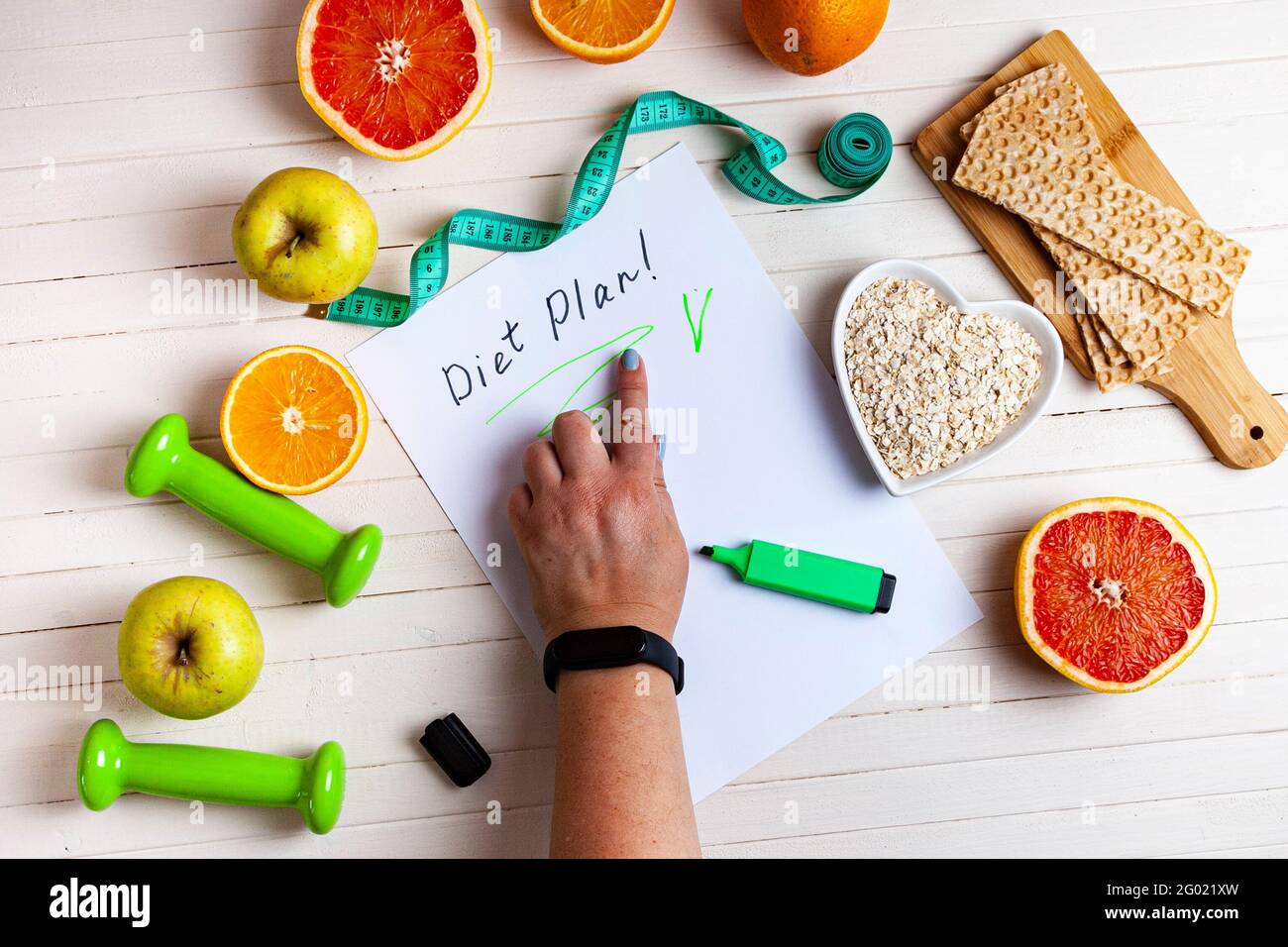 Weibliche Hand zeigt den Diätplan. Gesunde Ernährung, Kurzhanteln und Maßband auf einem Holzhintergrund. Stockfoto