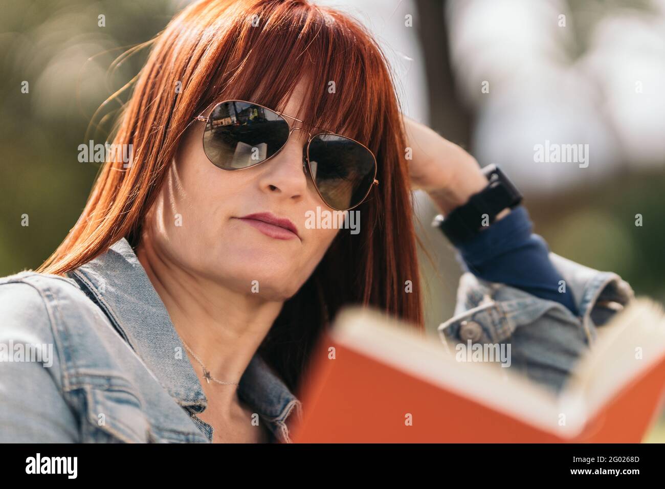 Eine rothaarige Frau mit Sonnenbrille liest ein Buch, das an einem sonnigen Tag seitlich auf einer Parkbank sitzt. Mittlere Nahaufnahme. Selektiver Fokus. Stockfoto
