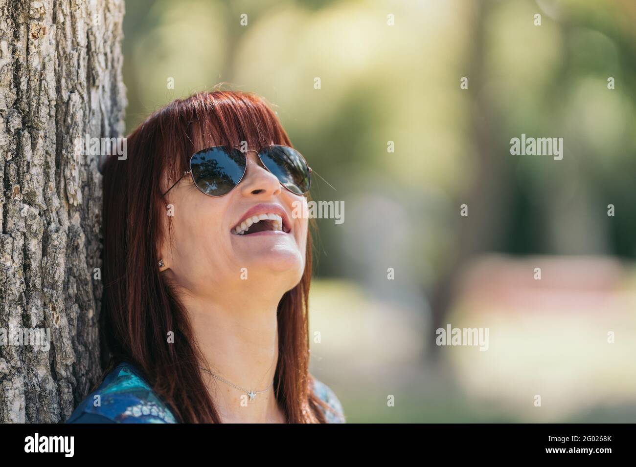 Eine rothaarige Frau mit Sonnenbrille lacht an einem sonnigen Tag gegen einen Baum. Mittlere Nahaufnahme. Selektiver Fokus. Stockfoto