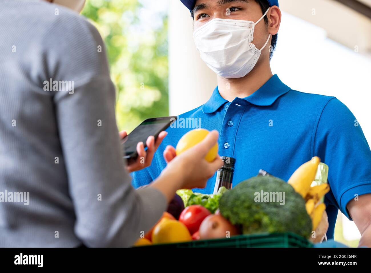 Hygienischer asiatischer Lieferer, der Gesichtsmaske trägt, während er Lebensmittel an den Kunden zu Hause liefert, und die Lieferung von Lebensmitteln in der Zeit des Pandemiekonzepts Stockfoto
