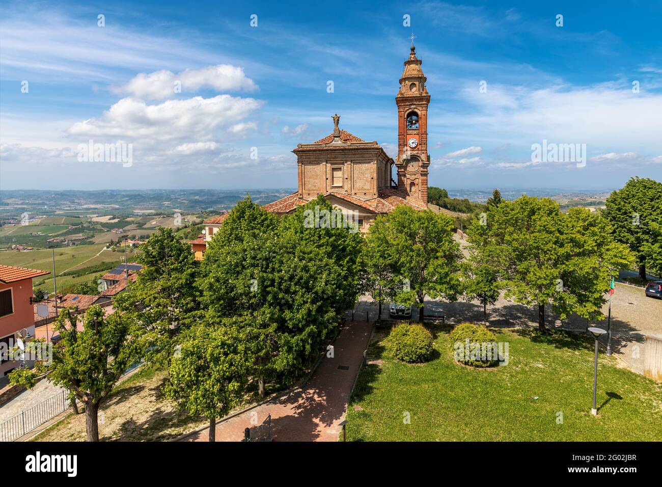 Blick auf die alte katholische Kirche unter grünen Bäumen unter schönem blauen Himmel mit weißen Wolken in der kleinen Stadt Diano d'Alba, Piemont, Norditalien. Stockfoto