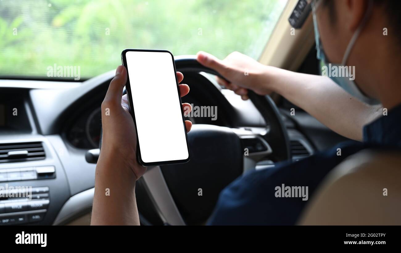 Kurzer Anschuss eines jungen Mannes, der eine Schutzmaske trägt und während des Autofahrens ein Smartphone verwendet. Stockfoto