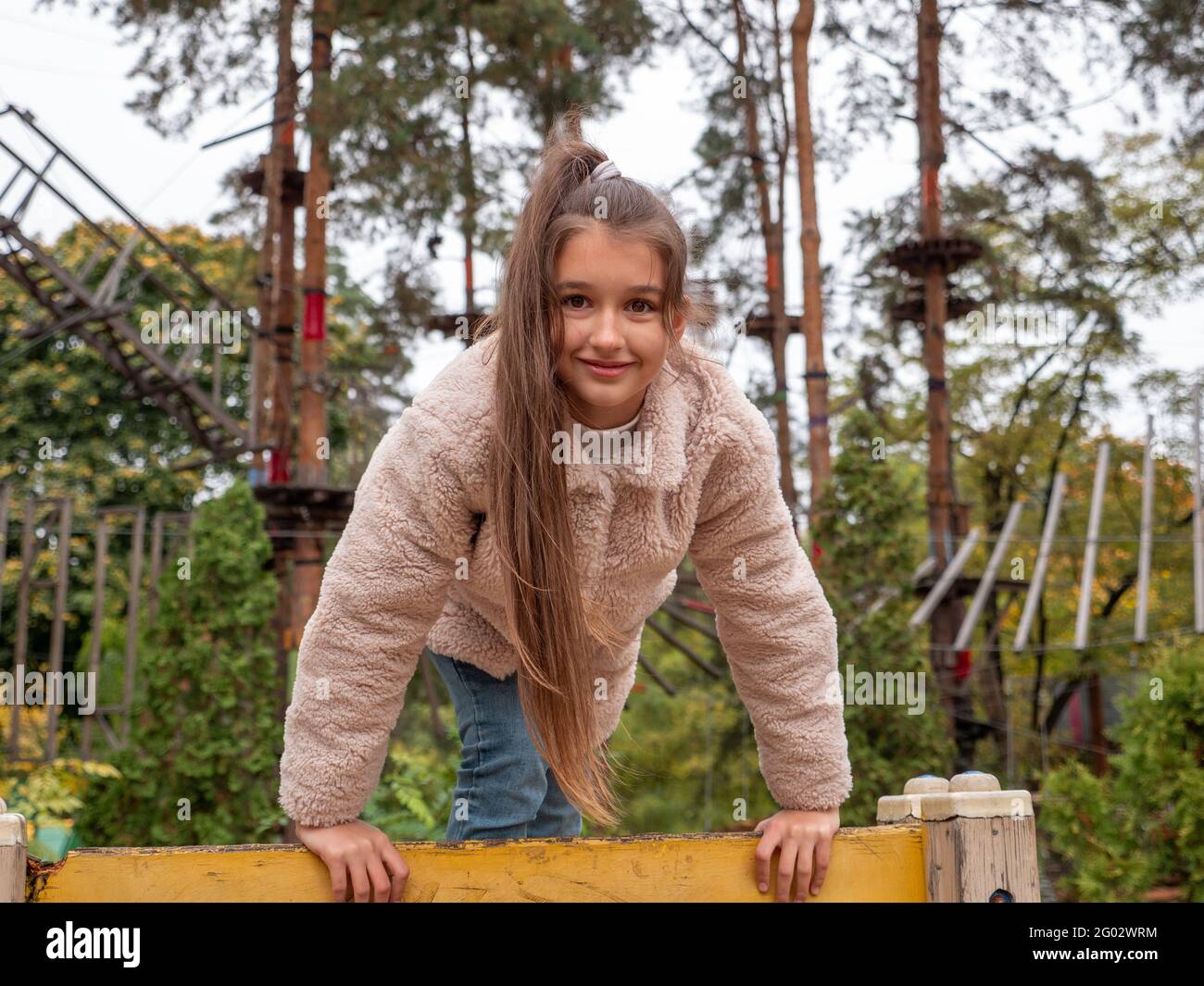 Glücklich lächelndes Teenager-Mädchen in einem Kunstpelzmantel und mit langem Pferdeschwanz klettern den gelben Kletterer auf dem Kinderspielplatz. Seilpark mit Pinien Stockfoto