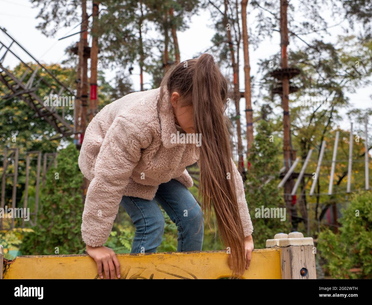 Glücklich lächelndes Teenager-Mädchen in einem Kunstpelzmantel und mit langem Pferdeschwanz klettern den gelben Kletterer auf dem Kinderspielplatz. Seilpark mit Pinien Stockfoto