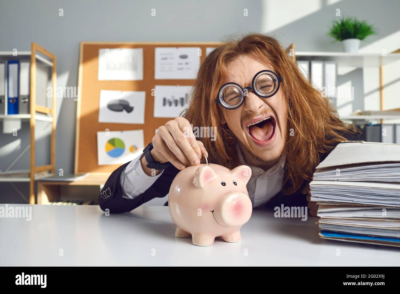 Glücklich verrückt aussehenden jungen Mann macht lustige Gesicht und Putting Münze in rosa Sparschwein Stockfoto