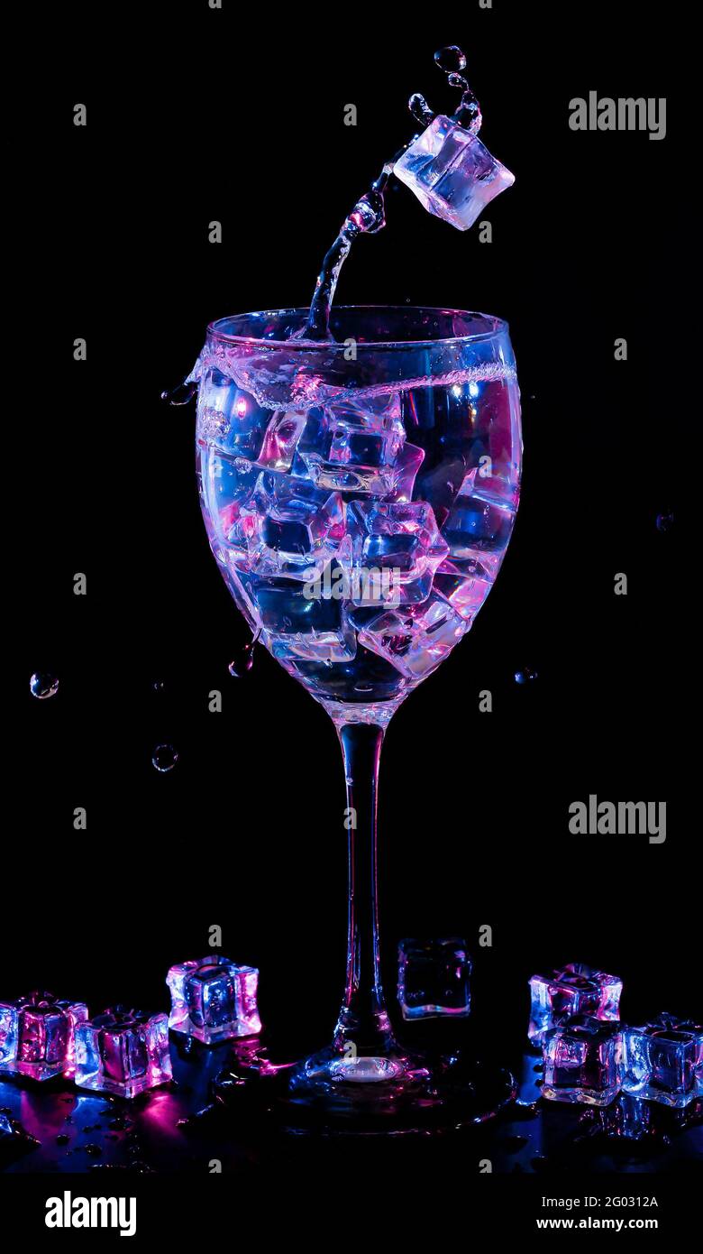 Eiswürfel, die mit Flüssigkeit in ein Weinglas fallen. Zweifarbige Hintergrundbeleuchtung. Stockfoto