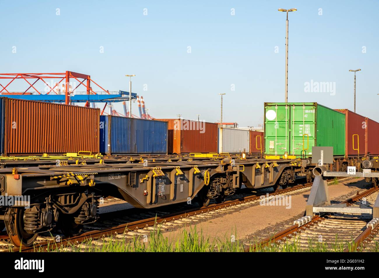 Eisenbahngüterwagen mit Containern auf Schienen im Hafen an einem sonnigen Tag mit blauem Himmel und Containerbrücken im Hintergrund Stockfoto