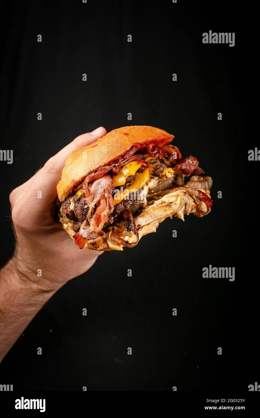 Hand ergreifend einen großen handgemachten Burger mit Käse, Speck, Zwiebeln, Tomaten und Cheddar. Stockfoto