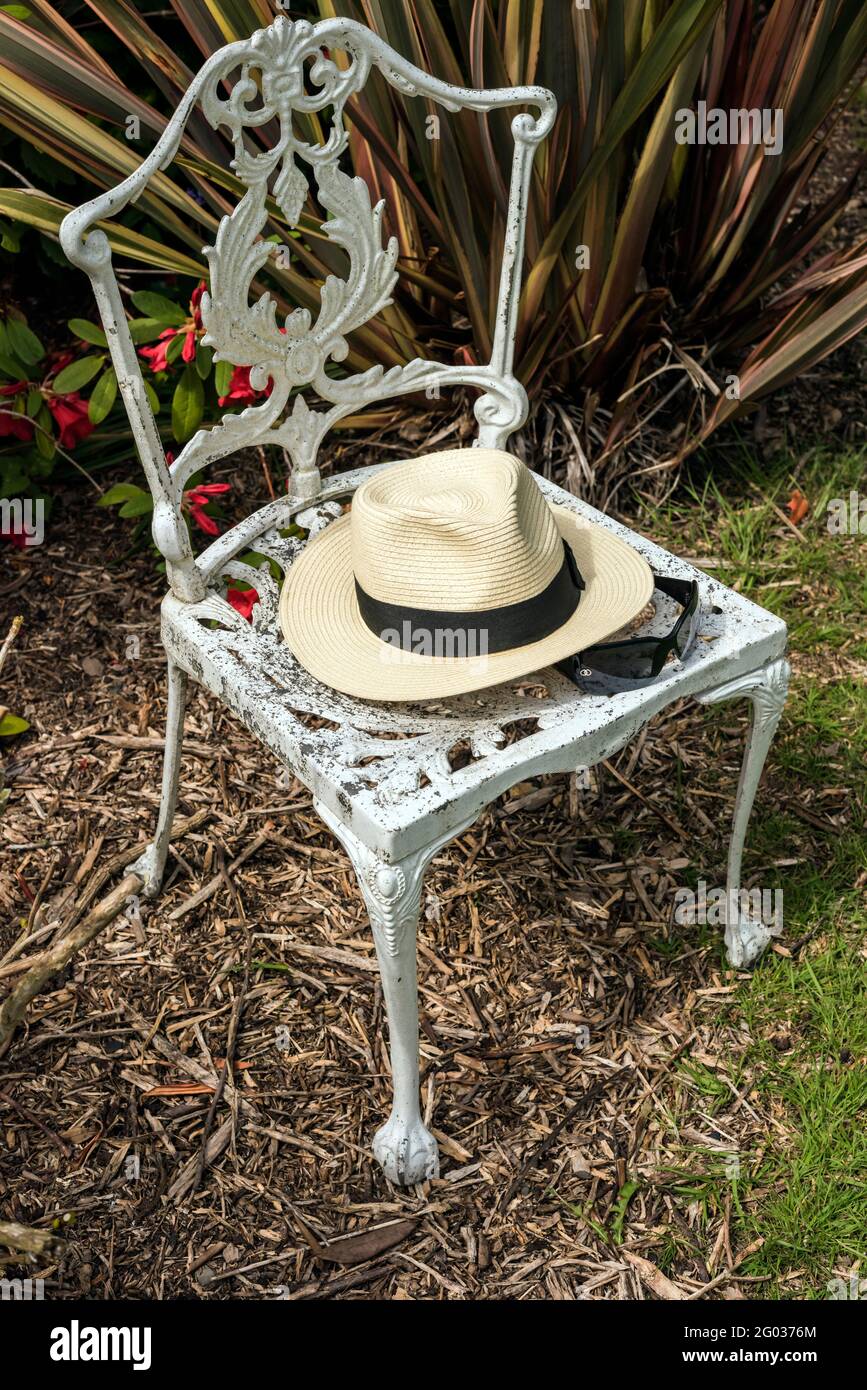 Panamahut und Sonnenbrille, platziert auf weißem Metallstuhl neben buntem Phormium und rotem Rhododendron. Stuhl auf Rinde und Gras gelegt. Konzept, Stockfoto