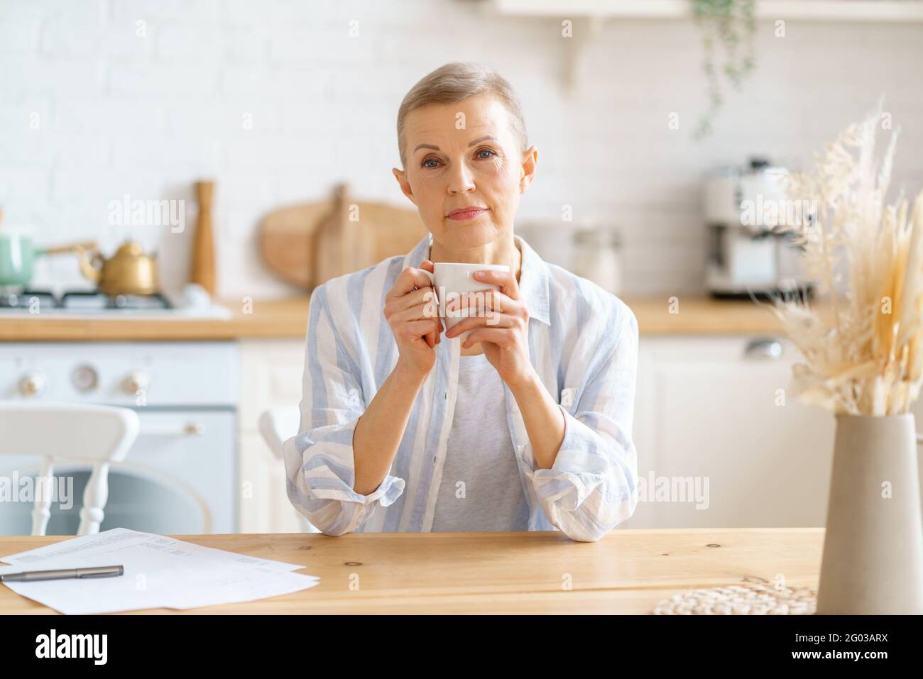 Morgengetränk. Schöne ältere Frau, die zur Kamera schaut und Kaffee oder Tee trinkt und dabei an etwas denkt, während sie am Holztisch in der Küche sitzt, selektiver Fokus auf Frau. Der Lebensstil der reifen Menschen Stockfoto