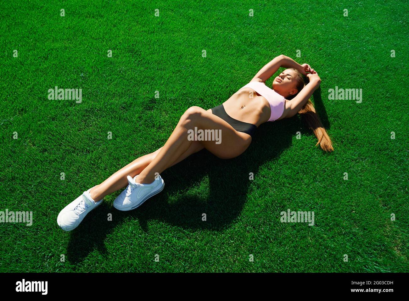 Draufsicht auf eine ziemlich junge muskulöse Läuferin, die auf frischem, hellgrünem Gras im Stadion liegt. Schöne Fitnesswoman mit Ruhe nach dem Training. Konzept des Trainings, gesunder Lebensstil. Stockfoto