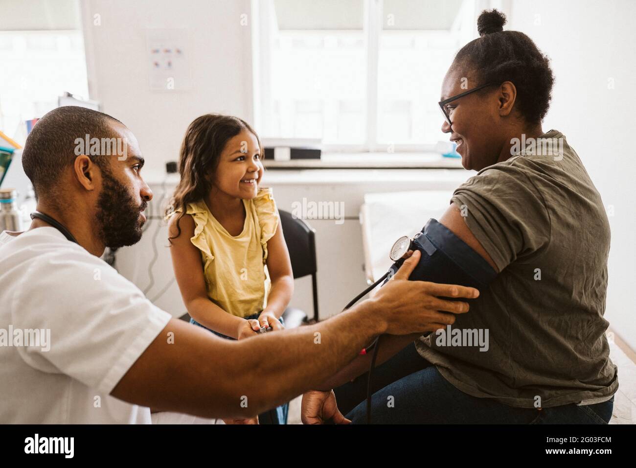 Lächelnde Mutter und Tochter, die einander anguckend, während ein männlicher Arzt den Blutdruck misst Stockfoto