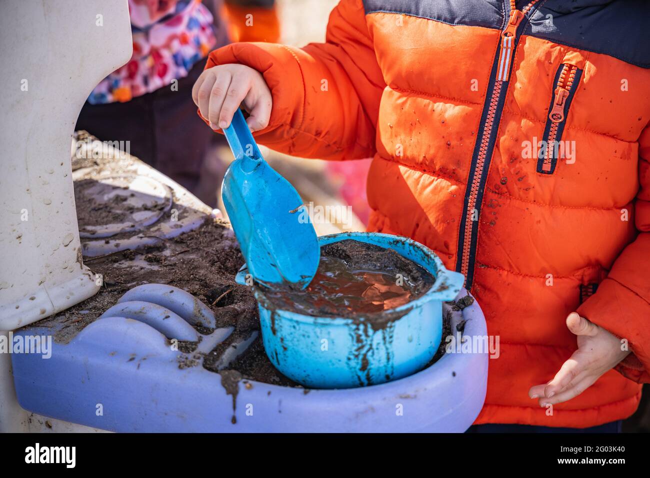 Nahaufnahme eines nicht erkennbaren jungen Kindergartenkindes in Winterkleidung und -Jacke, der mit einem blauen Plastikeimer und einer mit Schlamm gefüllten Schaufel spielt. Stockfoto